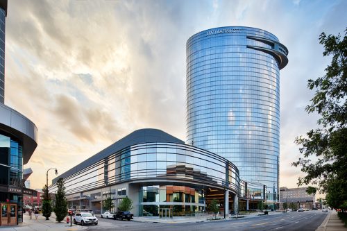 Street-level view of a curved glass podium and tall cylindrical tower with reflective glazing under a dramatic evening sky.