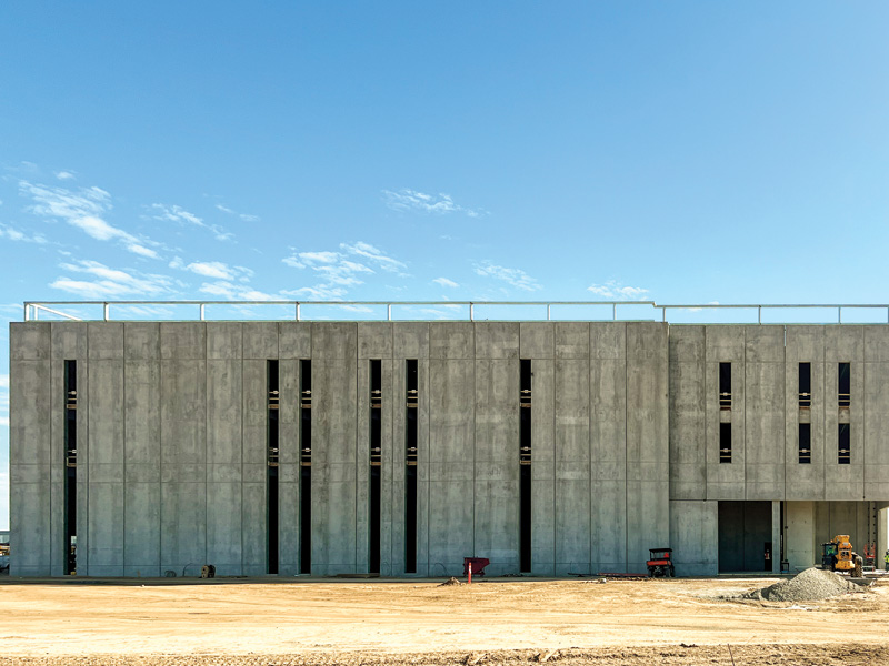 Construction site featuring a modern concrete building with vertical slits and equipment nearby against a clear blue sky.