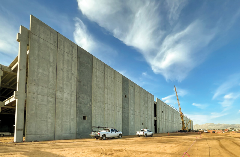 Construction site featuring a large concrete wall and machinery under a blue sky with white clouds, flanked by work vehicles.