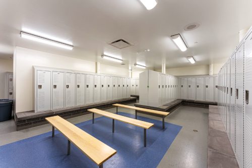A spacious, well-lit locker room featuring metal lockers, two wooden benches, and a blue mat on the floor.