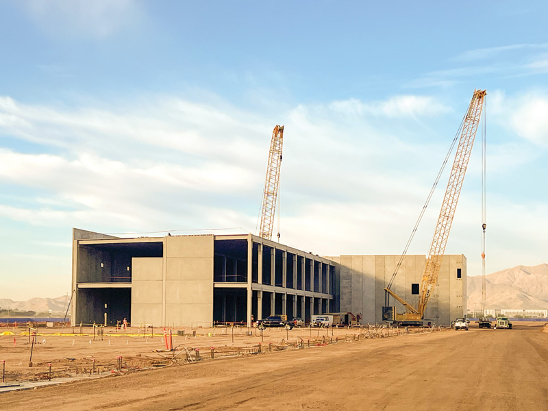 A concrete building under construction, equipped with two tall cranes, set against a sunny sky and distant mountains.