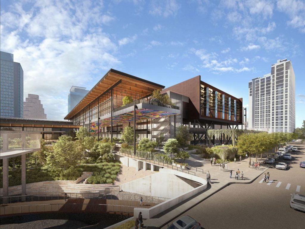 A modern building with a wooden roof, colorful artwork, and lush landscaping surrounded by city skyscrapers and pedestrians.