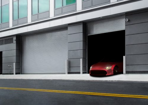 Red sports car exits through a high-performance rolling door on a modern building with sleek gray exterior and closed grilles.