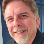 A close-up selfie of Tony Gambrall with wavy, light-colored hair in a dark shirt, against a blue wall background.
