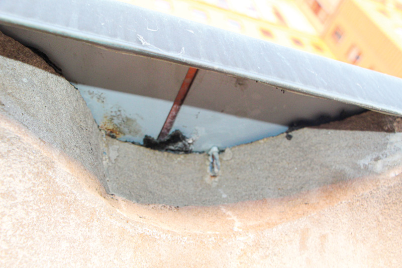 Close-up of a weathered building edge showing cracks, rusted metal, and possible water damage at the junction of roof and wall.