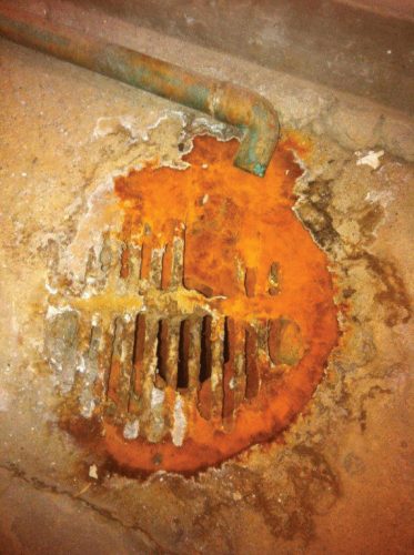 Rusty circular floor drain with slotted grate, surrounded by damaged concrete and a greenish pipe above.