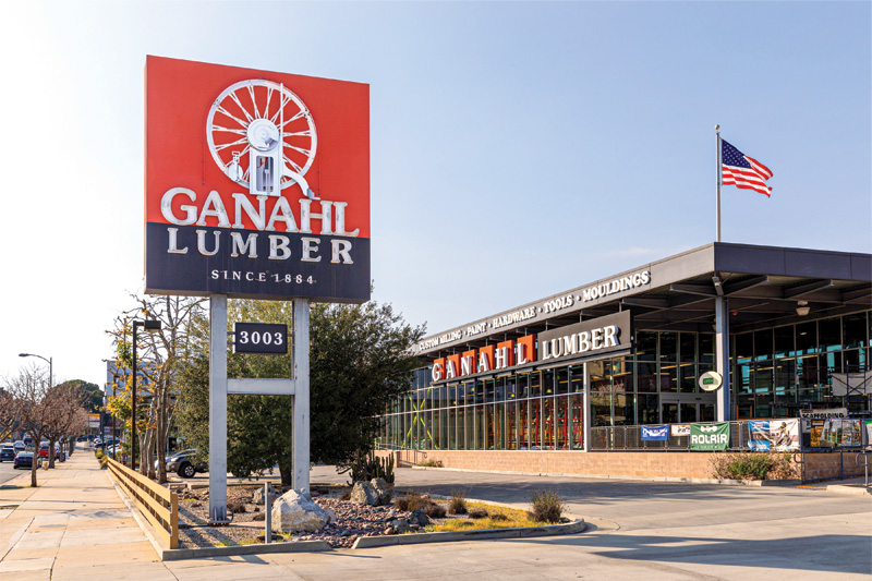 Exterior of Ganahl Lumber store with a prominent sign, U.S. flag, and clear blue sky. Established in 1884, it offers lumber and hardware.