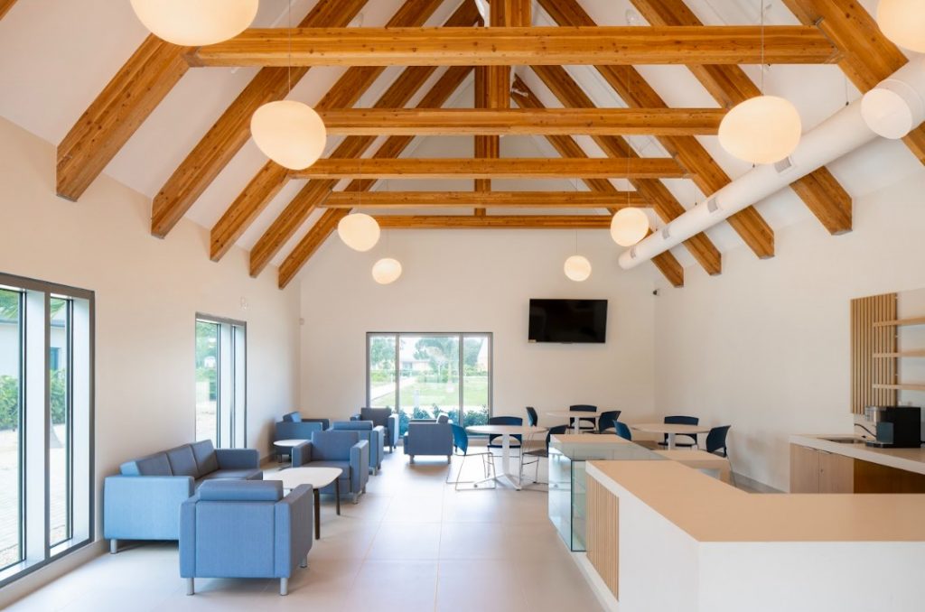 Bright, modern lounge with wooden beams, soft lighting, and gray-blue couches, featuring a bar area and large windows overlooking greenery.