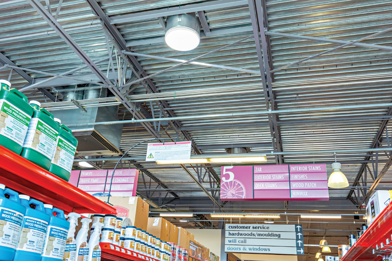 A view of a hardware store aisle with colorful cleaning products on shelves and overhead signs for various paint-related items.