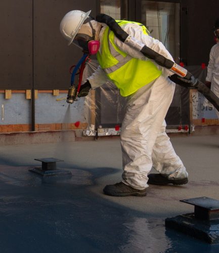 A construction worker in a yellow safety vest and protective gear uses a spray gun to apply material to a smooth, wet floor.