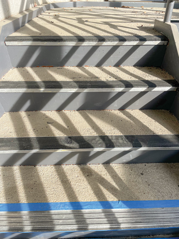 Close-up of concrete stairs with black rubber treads and diagonal shadows cast by railings, revealing a clean, textured surface.