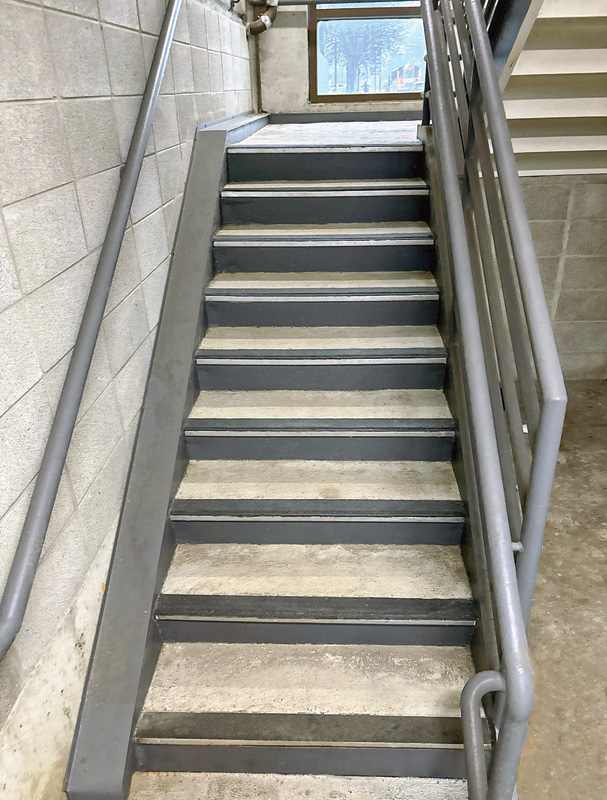 A view of a grey concrete staircase with metal railings, leading to a window at the top, surrounded by a plain wall.
