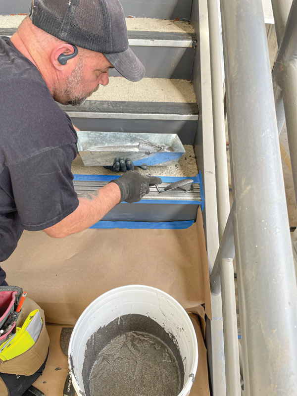 A worker applies a gray cement mixture to steps using a trowel, with a bucket of the mixture and protective tarp on the floor.