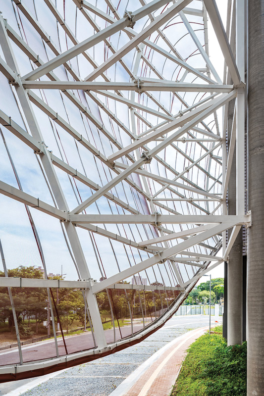 A close-up view of a modern building’s curved glass facade framed by white metal beams, with greenery and a path in the foreground.