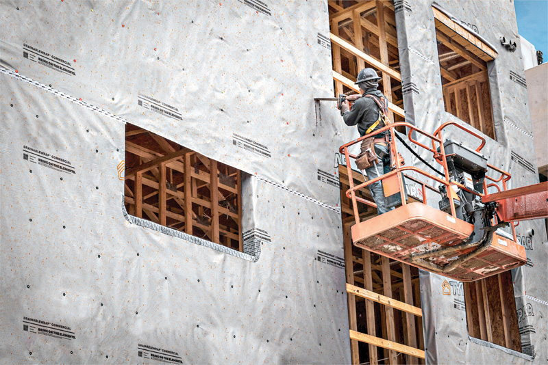 A construction worker in a safety helmet uses a tool from an elevated platform to work on a building wrapped in insulation material.