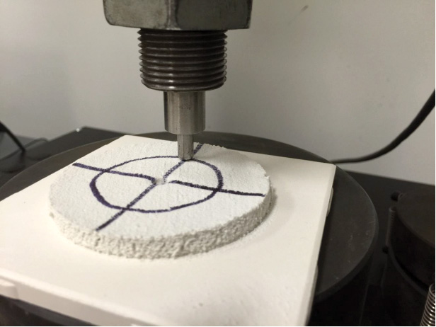 A close-up of a cylindrical compressive testing apparatus applying pressure to a white circular specimen marked with intersecting lines.