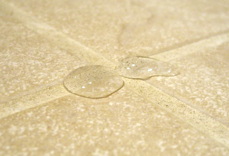 Two clear water droplets on a textured, light-colored tile surface, highlighting the shine and intricate pattern of the tile.