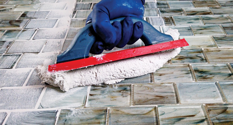 A gloved hand using a grout float to apply grout between glossy tiles on a floor, showcasing a tiling process in progress.