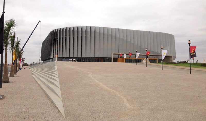 A sleek, modern sports arena with a curved exterior, set against a cloudy sky and flanked by decorative flags and palm trees.