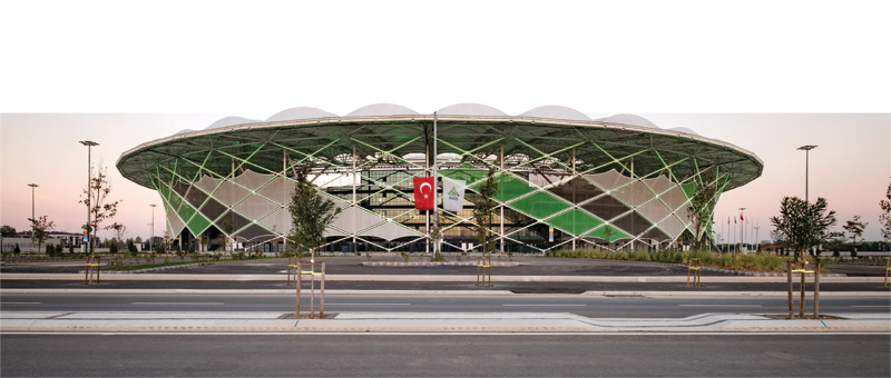 Modern stadium with a futuristic design, featuring green accents and surrounded by trees, located on a well-maintained street.