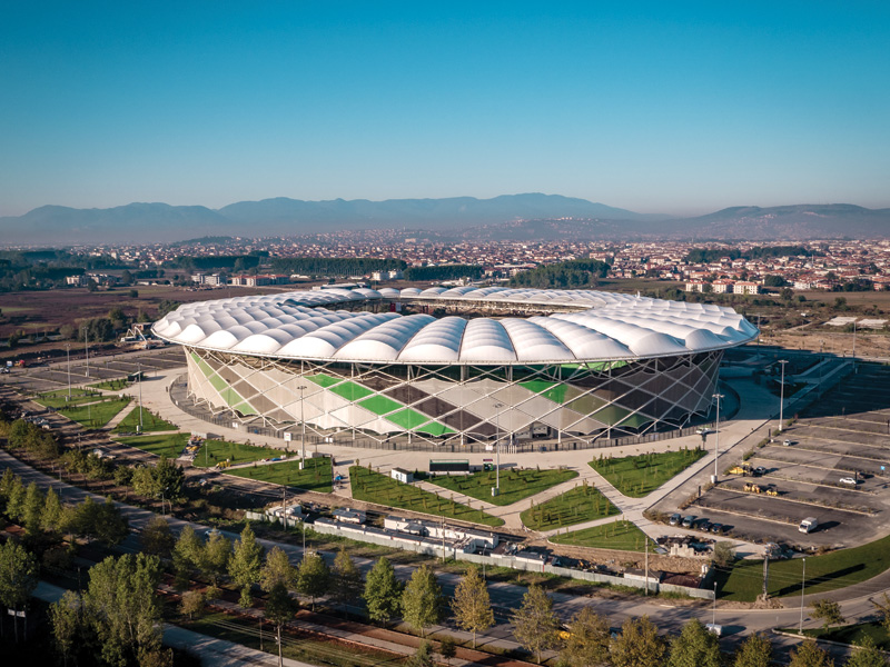 Aerial view of a modern stadium with a unique roof design, surrounded by green landscaping and distant mountains under a clear sky.