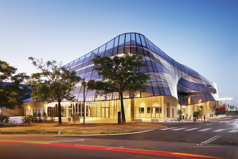 Modern building with a sleek, glass facade, surrounded by trees, illuminated at dusk with a gentle blue sky above.