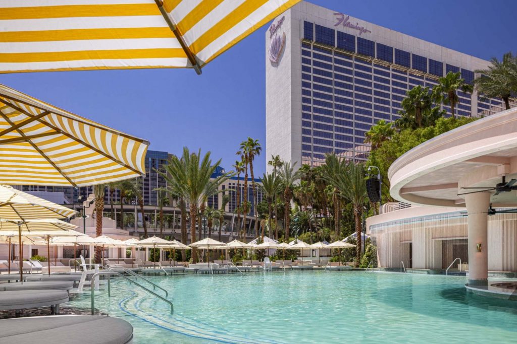 A vibrant pool surrounded by palm trees and striped umbrellas, with the Flamingo hotel towering in the background under a clear blue sky.