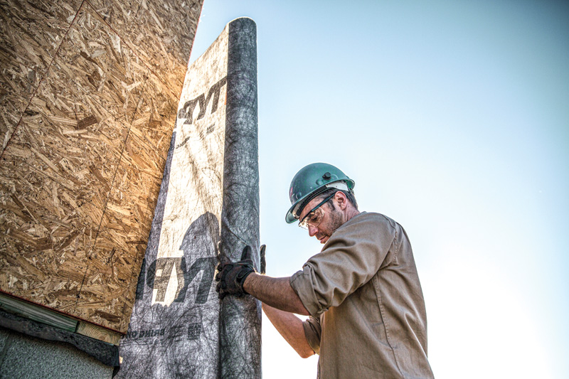A construction worker, wearing a helmet and gloves, installs a roll of building material against a wooden structure under a clear sky.