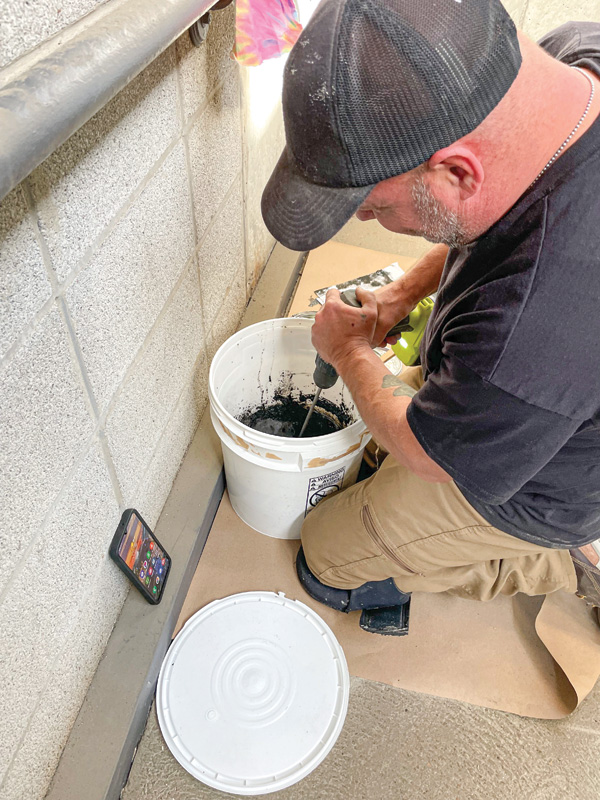 A person kneels beside a bucket mixing black material with a drill, while a smartphone rests on the ground nearby.