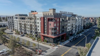 A modern multi-story apartment building named "Bilaik Station" with red and gray accents, surrounded by trees and parked cars.