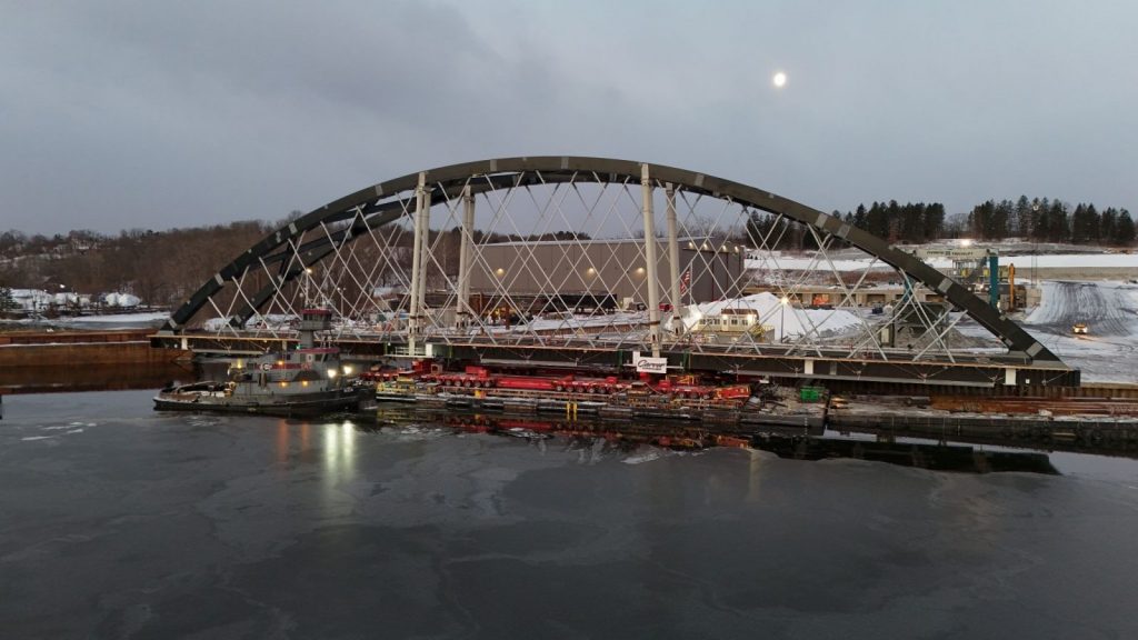 A tugboat docks near an arched bridge over icy water, with industrial buildings and a full moon in the background on a winter evening.