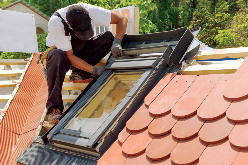 A worker installs a skylight on a sloped roof, surrounded by red roof tiles and green trees in the background.