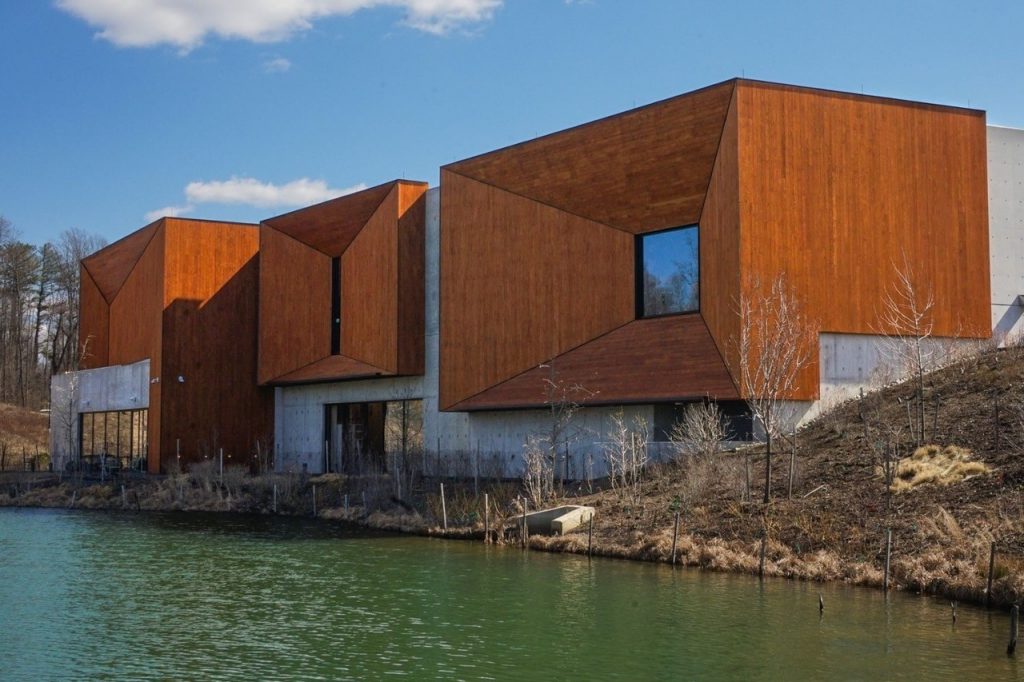 Modern angular architecture featuring rust-colored façades and large windows, set beside a serene green lake against a blue sky.