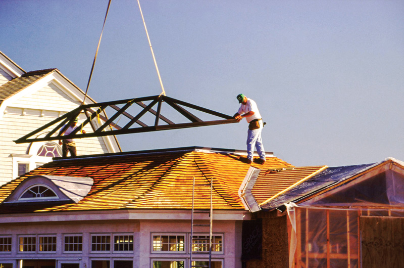 Two construction workers install a roof truss on a sloped roof under clear blue skies, with tools and equipment nearby.