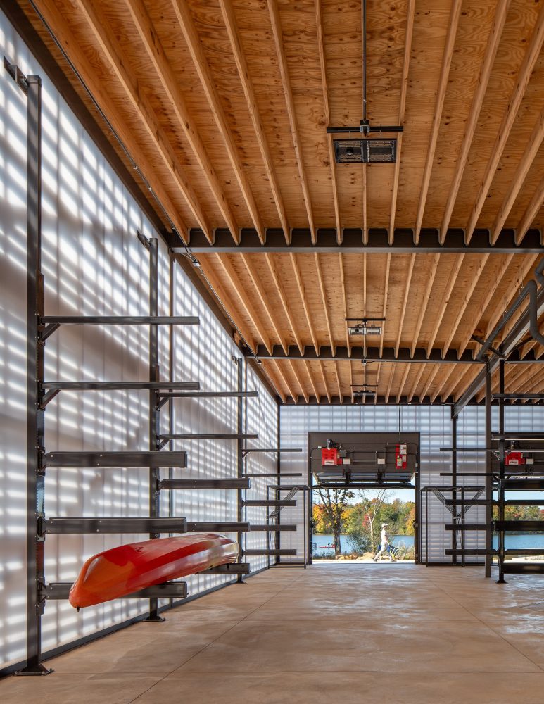 Interior of a modern boathouse with wooden beams, metal shelving, and a red kayak, leading out to a scenic view of water and trees.