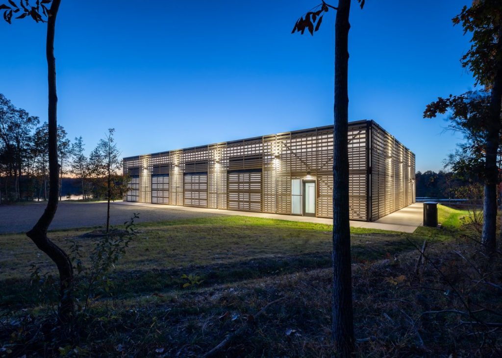 Modern wooden building illuminated at dusk, surrounded by trees, with a clear blue sky and water in the background.