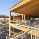 A partially constructed building showcasing wooden beams, spacious open levels, and safety railings against a clear blue sky.