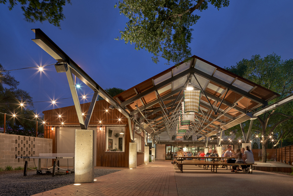 Modern outdoor dining space features a spacious pavilion with string lights, surrounded by trees, and buzzing with lively diners.