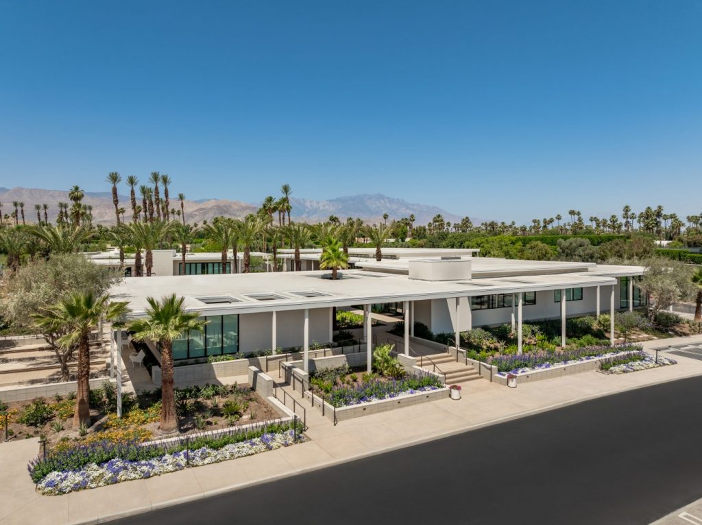 Modern building surrounded by lush palm trees and colorful landscaping, set against a clear blue sky and mountain backdrop.