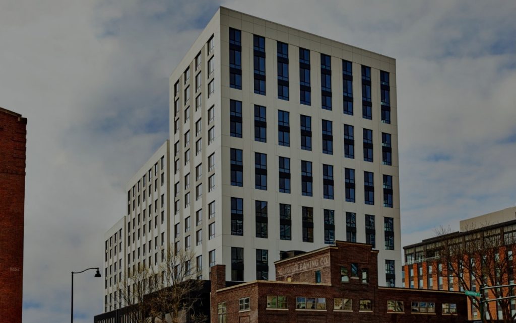 A modern, white multi-story building towers over an older brick structure, with a cloudy sky in the background.