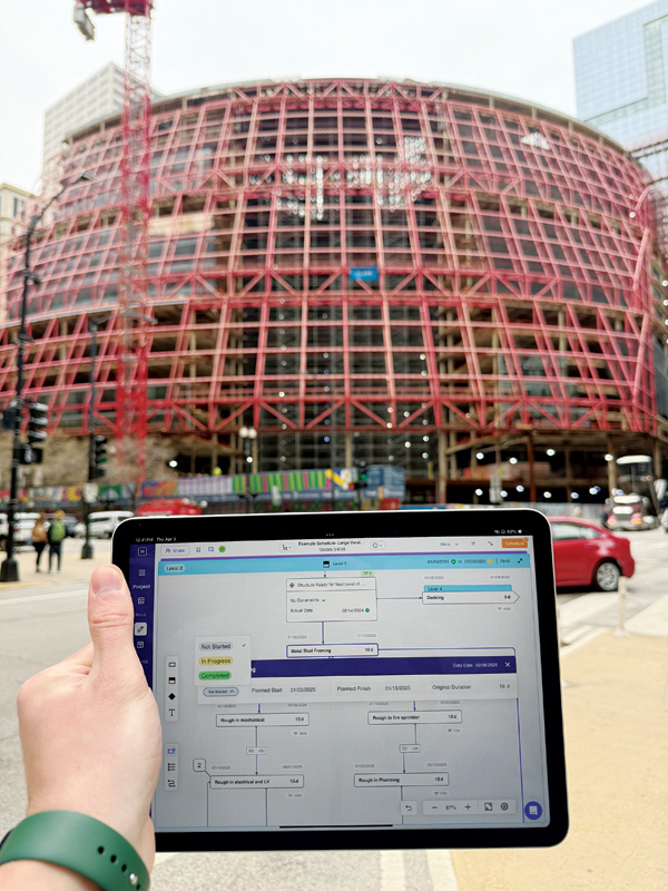 A hand holds a tablet displaying project details in front of a large, partially constructed building with a red frame.