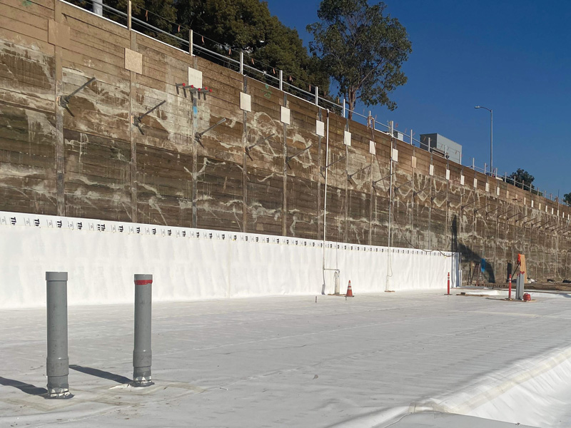 Construction site featuring a large, partly concrete wall with white sheeting below, red and gray pipes, and a clear blue sky.