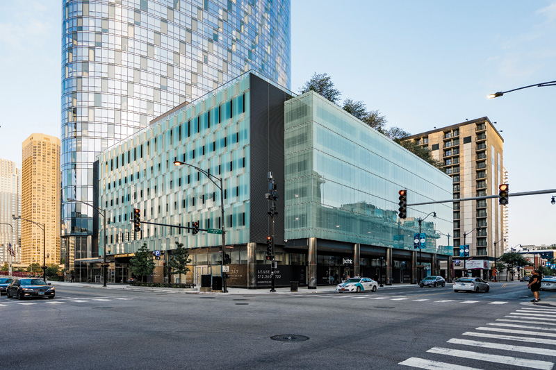 Modern buildings at a bustling intersection, showcasing glass and brick facades against a clear sky. Traffic signals and parked cars visible.