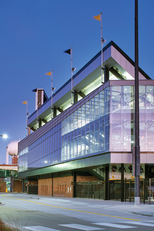 Modern stadium building with large glass windows, vibrant flags, and illuminated interior, set against a twilight sky.