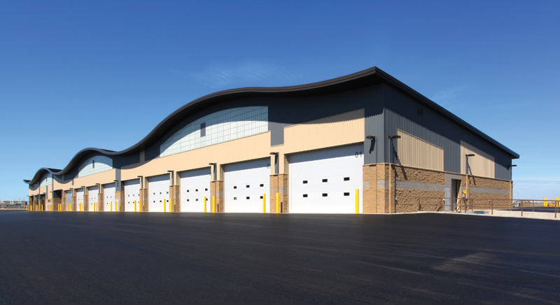 A modern warehouse with a wavy roof and multiple loading bay doors, set against a clear blue sky and smooth asphalt pavement.