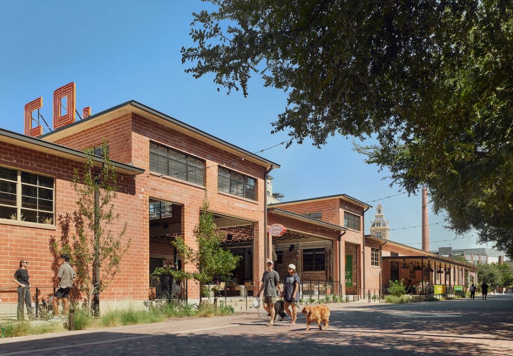 Two people walk dogs along a brick pathway lined with green plants, with a historic red-brick building and blue sky in the background.