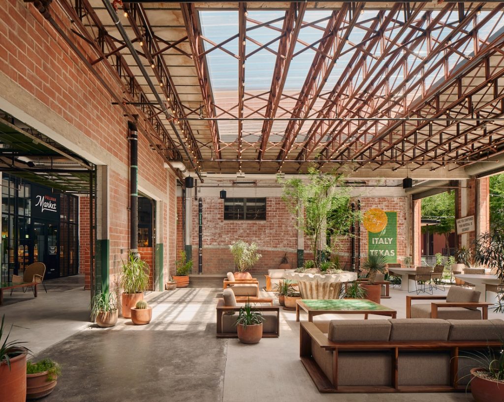 A modern, spacious indoor market with industrial decor, lush plants, and cozy seating, illuminated by natural light from a skylight.