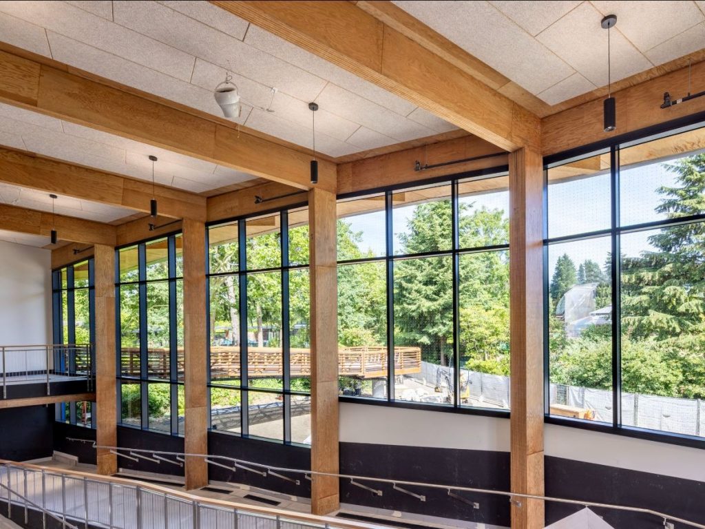 Bright, modern interior with large windows, wooden beams, and a view of green trees outside. Minimalist design with an inviting atmosphere.