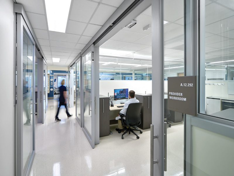 A modern healthcare corridor with glass-walled offices; a person walks by while another sits at a desk in the provider workroom.
