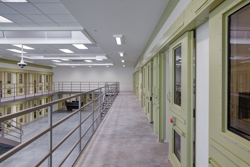 A view of a modern prison corridor featuring green inmate doors, metal railings, and bright overhead lighting.